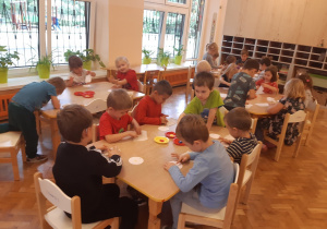 Dzieci z grupy pomarańczowej siedzą przy stolikach. Za pomocą palców maczanych w farbie: żółtej, niebieskiej i czerwonej malują na białych, papierowych kołach - wykonują swój medal przedszkolaka.